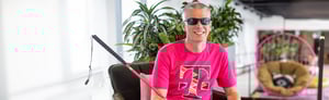 A young blond man is seated, and smiles as he holds a magenta cane across his lap. He wearing dark glasses and a magenta T logo shirt.