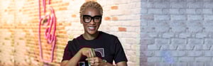 A young Black woman with blond hair and striking glasses wears a T logo shirt and stands in front of a weathered brick wall while signing
