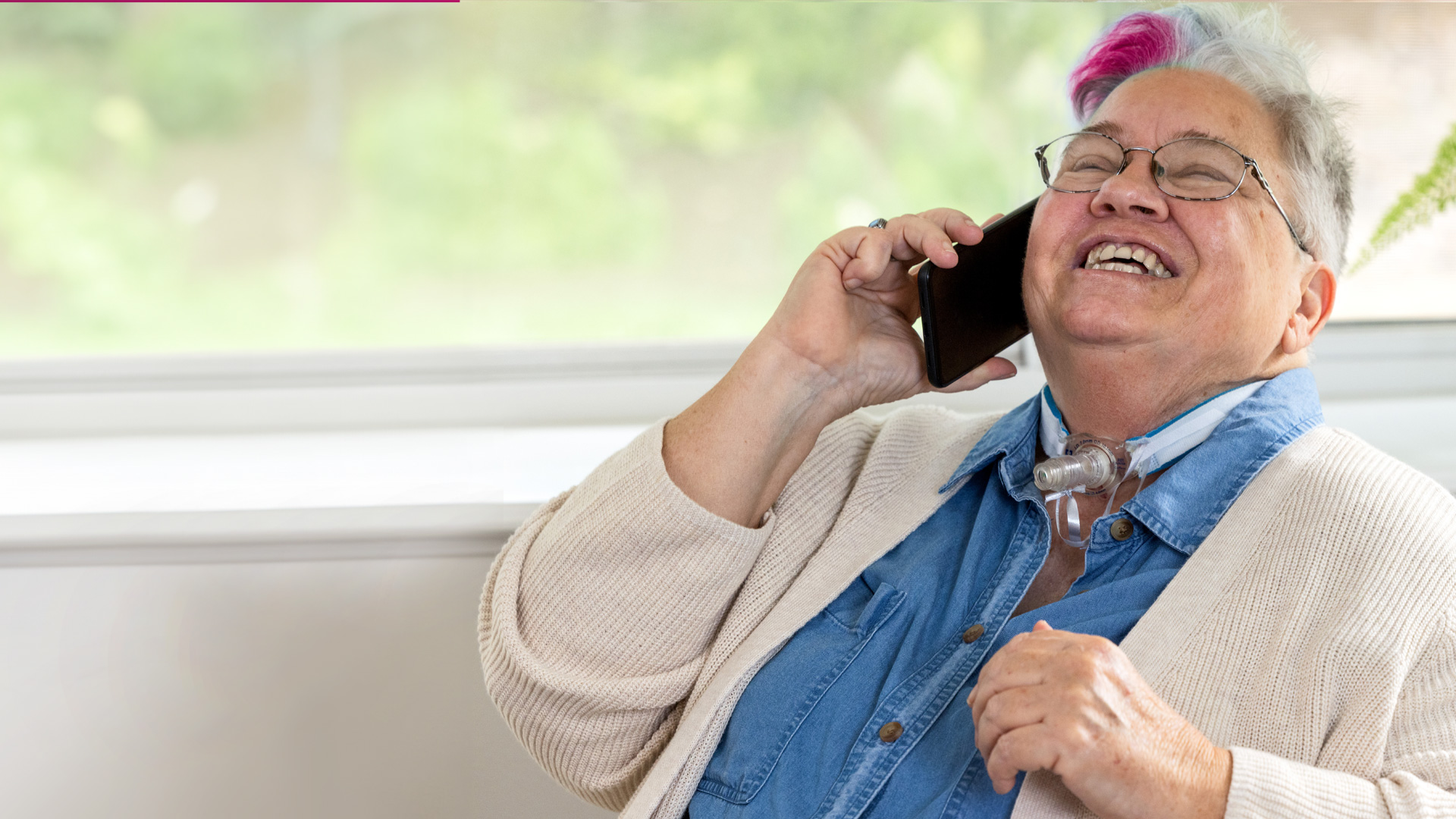 An older woman with a phone to her ear smiles with joy. She has a swath of magenta in her gray hair and a visible tracheostomy tube.
